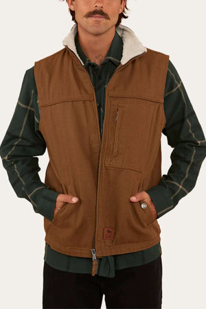 A man with a mustache wears the Ringers Western High Country Vest, featuring cotton canvas fabric and a fleece collar, over a dark green plaid shirt. He stands with hands in vest pockets against a plain off-white background.