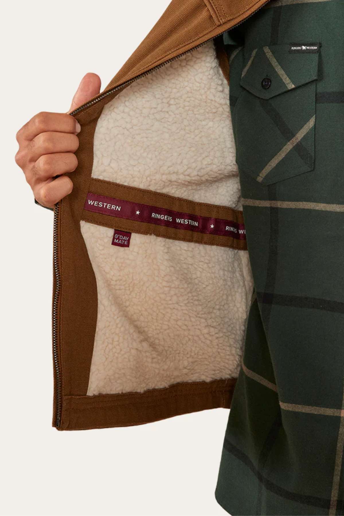 A person holds open a brown Ringers Western High Country Vest to show its soft white fleece lining and maroon “WESTERN RINGERS” label, worn over a green and black plaid shirt.