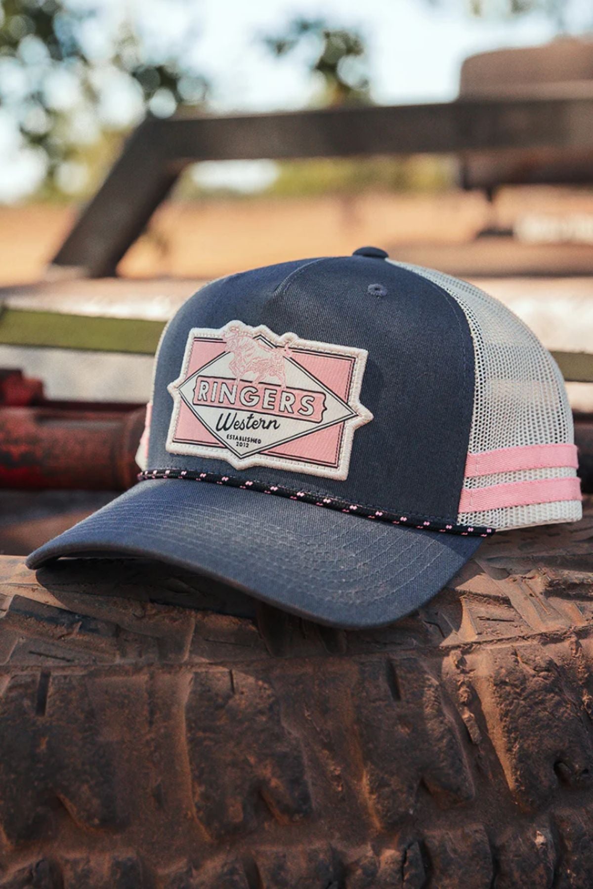 A Ringers Western Hometown Trucker Cap in navy blue and white with a pink patch rests on a dusty tire outdoors, featuring an adjustable snapback and blurred trees and equipment in the background.