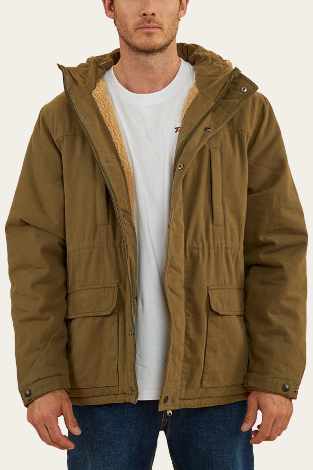 A man wearing a khaki Ringers Western Judd Jacket over a white t-shirt and blue jeans. The jacket, featuring large front pockets and a sherpa lining visible around the collar, has a removable hood. The man is standing facing the camera with his hands relaxed at his sides.