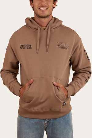 A man wears the Ringers Western KNX Oversized Hoodie in light brown, featuring "RINGERS WESTERN" and "RINGERS" text on the chest, made from soft brushed fleece. He stands with hands in the front pocket, paired with light blue jeans.