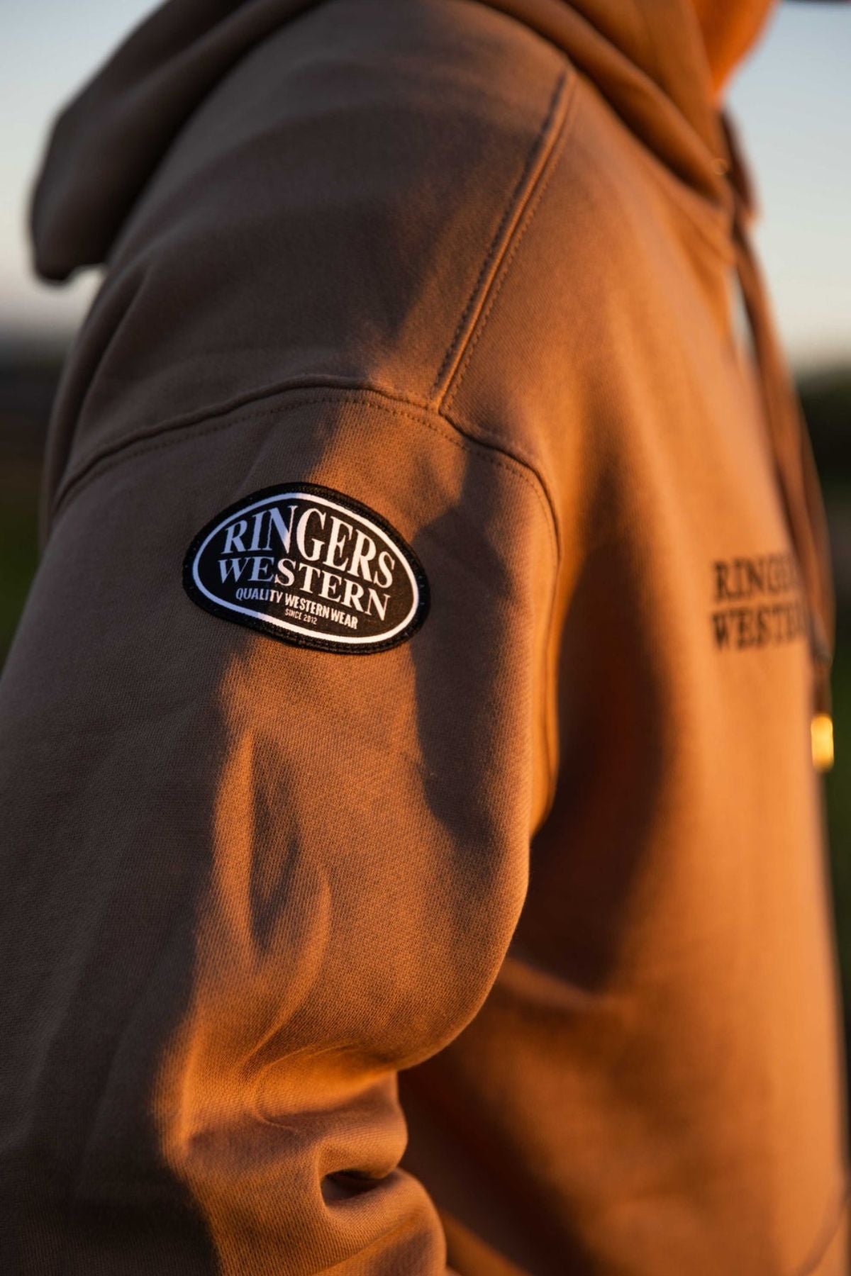 A close-up of a person wearing the Ringers Western KNX Oversized Hoodie, featuring "RINGERS WESTERN" on a sleeve patch and chest print, crafted from soft brushed fleece and shown in warm sunlight.