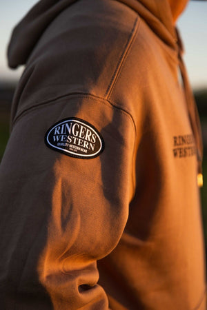A close-up of a person wearing the Ringers Western KNX Oversized Hoodie, featuring "RINGERS WESTERN" on a sleeve patch and chest print, crafted from soft brushed fleece and shown in warm sunlight.