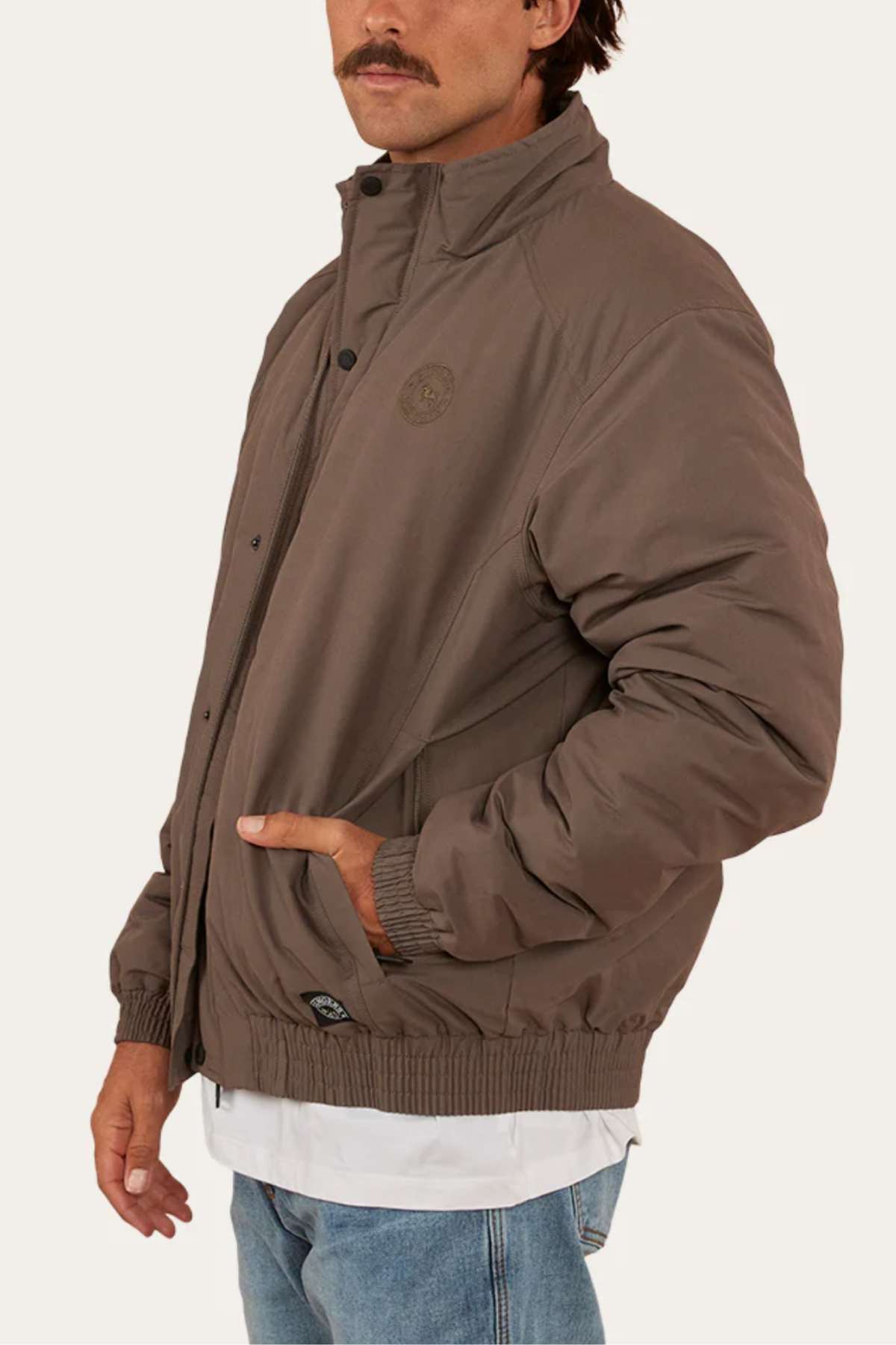 A man wearing a Ringers Western Kidman Jacket with a high collar, zipper front, and quilted lining stands with his left hand in the pocket of his jeans. Sporting a mustache, the jacket features a small circular logo on the left chest area. The background is plain light grey, ideal for an outdoor adventure.