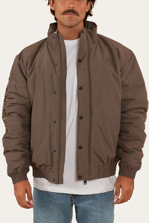 A person stands facing forward wearing a brown, high-collar, button-up Ringers Western Kidman Jacket over a white T-shirt. The jacket, perfect for an outdoor adventure, features elastic cuffs and a subtle emblem on the left chest. The person is also wearing light blue jeans and has short, dark hair and a mustache.
