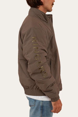 A person wears a brown Ringers Western Kidman Jacket with "RINGERS" written vertically on the sleeve. The jacket, perfect for outdoor adventures, features a stand-up collar. The individual is also dressed in blue jeans and a white shirt, standing sideways with their face partially visible.
