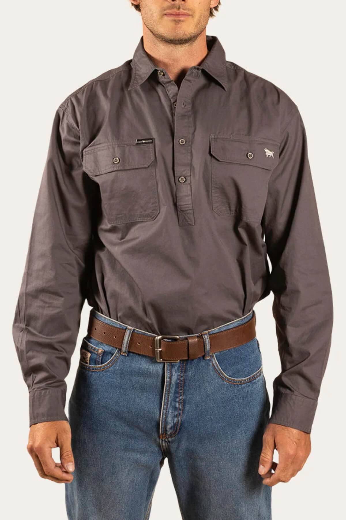 Man wearing a dark gray Ringers Western King River Half Button Shirt with two chest pockets and blue jeans, finished with a brown leather belt. The background is plain and light-colored.