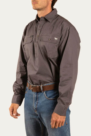Someone wearing a Ringers Western King River Half Button Shirt in dark gray with long sleeves and two chest pockets, paired with blue jeans and a brown belt, stands against a plain light background.