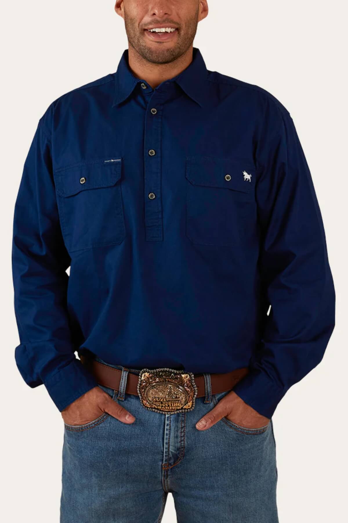 A person is standing with hands in their pockets, wearing a dark blue Ringers Western King River Half Button Shirt with two chest pockets, light blue jeans, and a brown belt with an ornate, large silver buckle. The background is plain and light-colored.