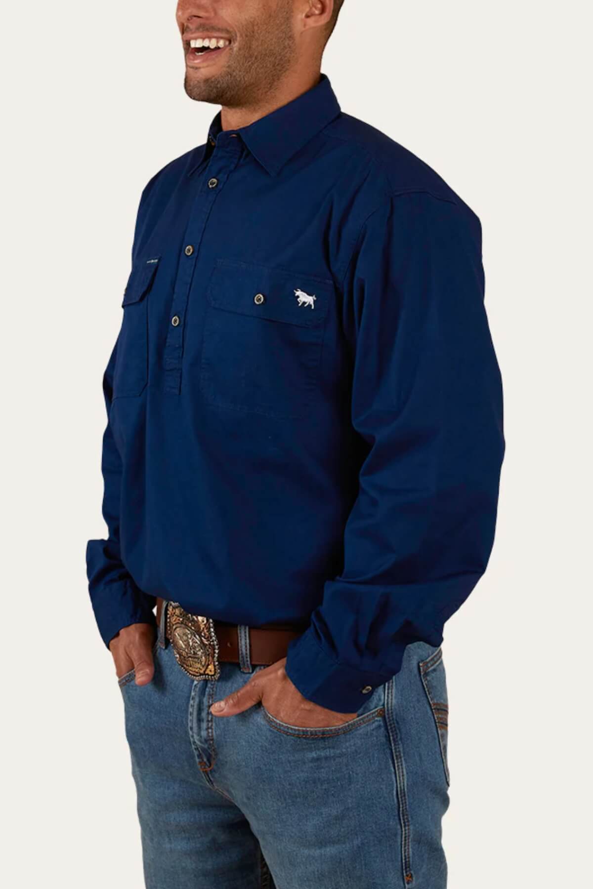 A person is wearing a dark blue, long-sleeved Ringers Western King River Half Button Shirt with buttoned pockets on the chest. The 100% cotton shirt features a small embroidered logo on the left pocket. They are also wearing blue jeans and a brown belt with an ornate buckle. They are standing and smiling.