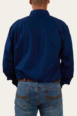 A person wearing a dark blue Ringers Western King River Half Button 100% cotton long-sleeve work shirt and blue jeans is shown from the back. The shirt is untucked, and the jeans have visible stitching on the back pockets. The person's hands are in their pockets, against a plain, light background.