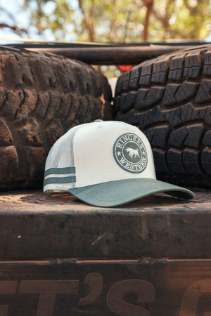 A Ringers Western McCoy Trucker Cap in light gray and green, made from durable cotton twill, sits on a dusty surface outdoors before two large mud-covered tires.