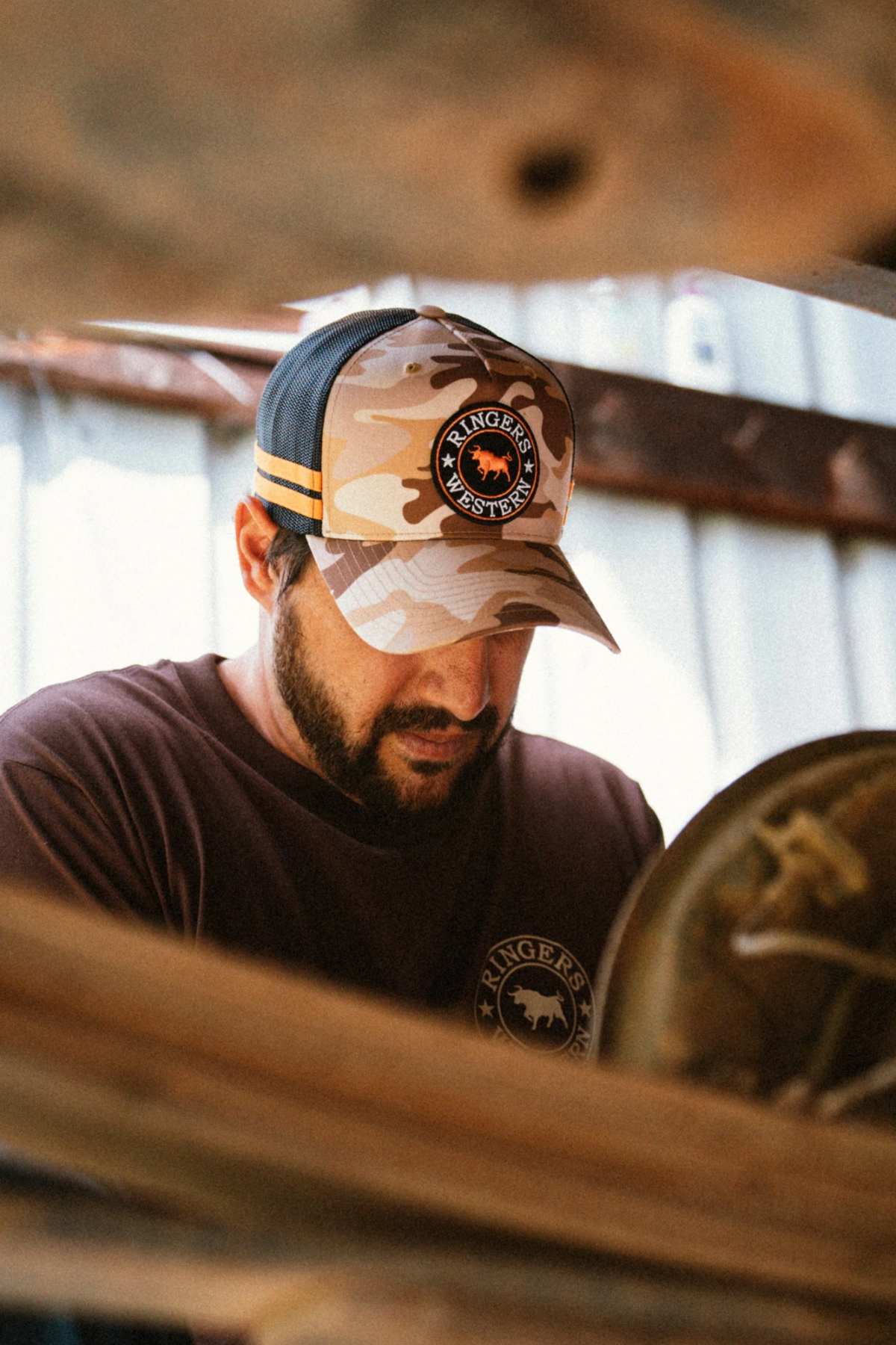 A man wearing a camouflage Ringers Western McCoy Trucker Cap with mesh back and logo patch, paired with a brown T-shirt, looks downward while working indoors near wooden beams.