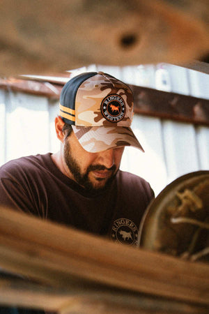 A man wearing a camouflage Ringers Western McCoy Trucker Cap with mesh back and logo patch, paired with a brown T-shirt, looks downward while working indoors near wooden beams.