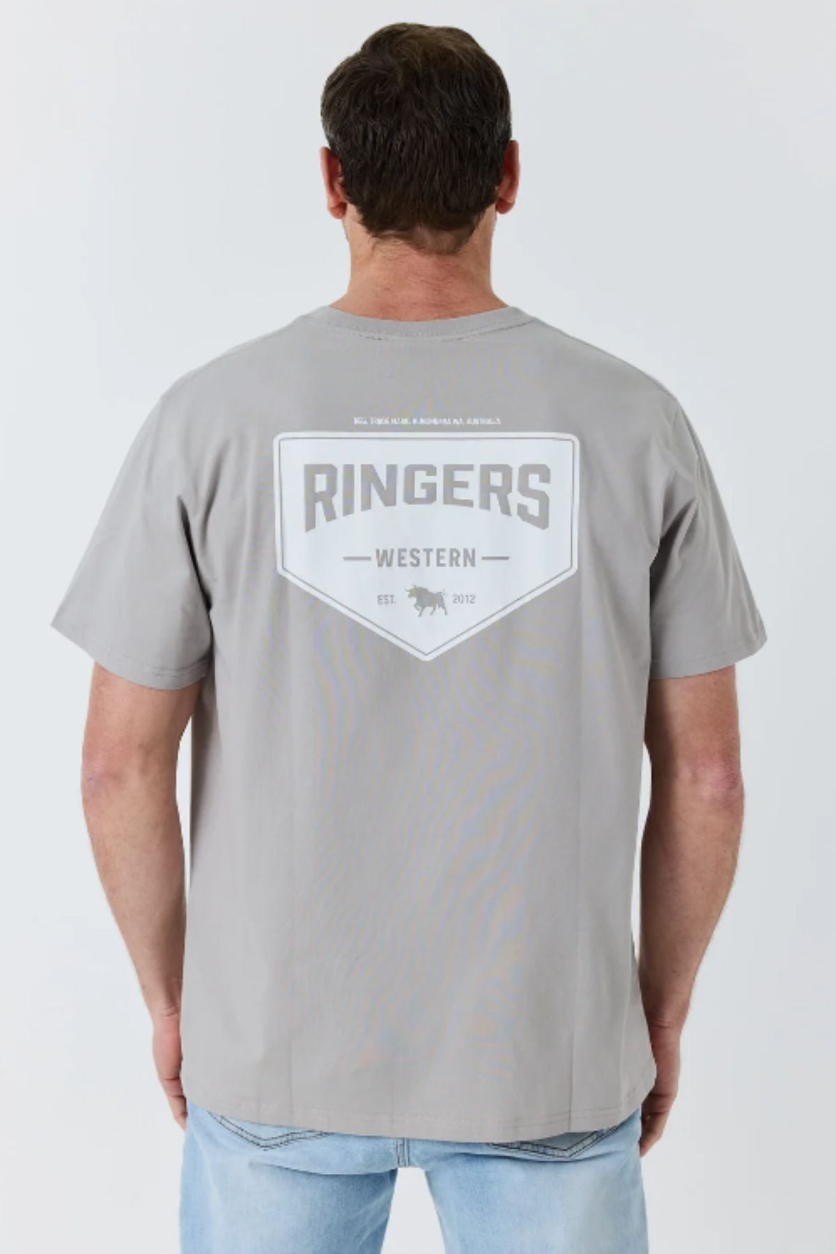 A man is wearing the Ringers Western New Squadron Loose Fit Tee, made from 100% cotton in light gray. The back of the shirt features “RINGERS WESTERN” in bold letters, along with a small bull head graphic and the text “EST 2012.” He is facing away from the camera.