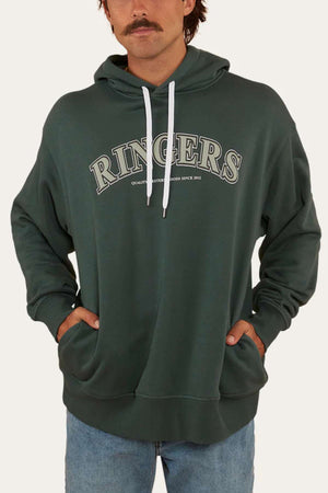 A person wearing an oversized dark green Ringers Western Princeton Hoodie with "RINGERS" printed across the chest in large white letters. The brushed fleece hoodie has hands in the front pockets and is paired with light blue jeans. The background is plain and off-white.