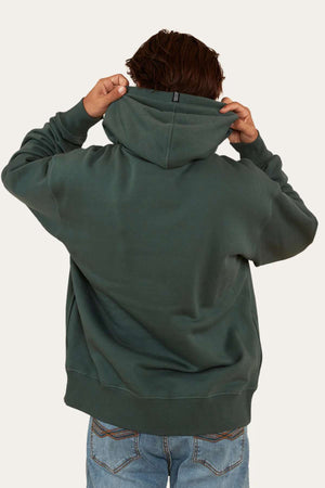 A person with short, brown hair is facing away and adjusting the hood of their dark green Ringers Western Princeton Hoodie. Made from brushed fleece and featuring an oversized fit, this comfortable hoodie pairs perfectly with their light blue jeans against a plain, light-colored background.