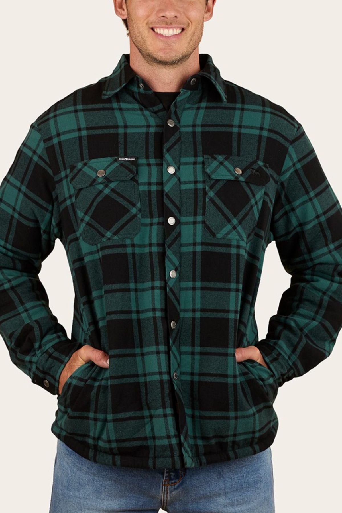 A person wearing the Ringers Western Sawyer Flannel Shirt in green and black plaid, featuring front pockets, smiles with hands in pockets. They are also dressed in blue jeans.
