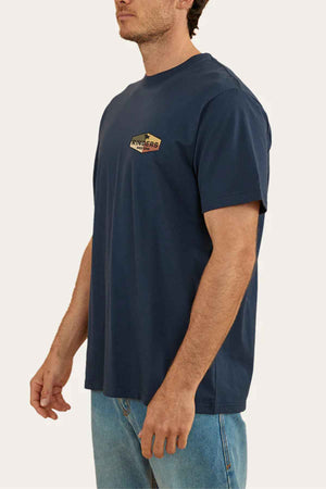 A man dressed in a dark blue Ringers Western Servo Loose Fit T-Shirt featuring a small chest logo, paired with light blue jeans, is standing sideways against a plain backdrop.