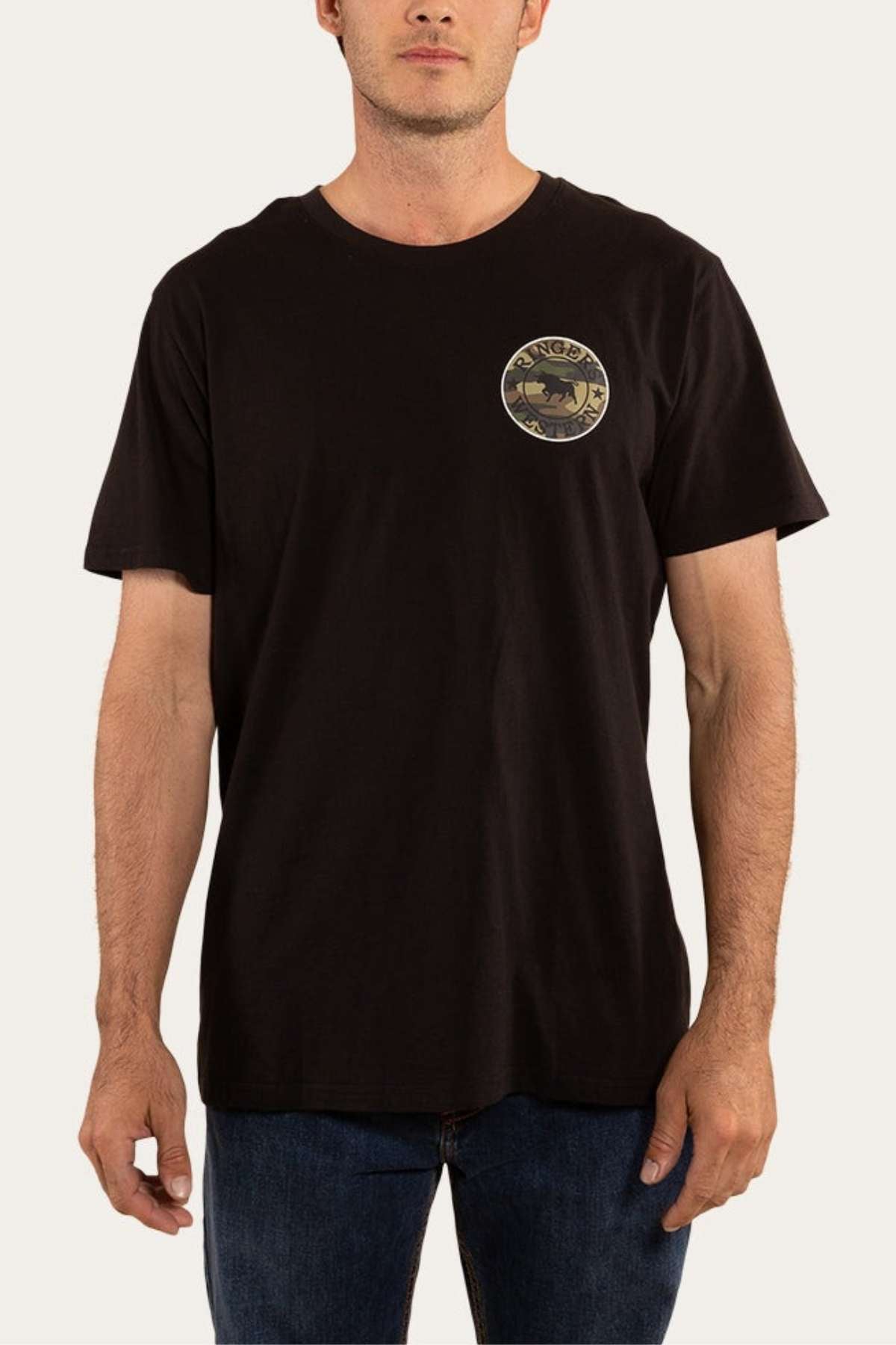 A person wearing the Ringers Western Signature Bull Classic T-Shirt—a black, comfortable short sleeve tee with a small circular logo on the left chest featuring an illustration and text. The classic fit tee is paired with blue jeans, set against a plain off-white background.