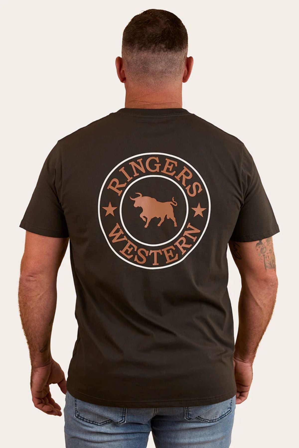 A man wears the Ringers Western Signature Bull Loose Fit T-Shirt made from 100% cotton, featuring a circular bull and stars design on the back, paired with light blue jeans while standing with arms relaxed.