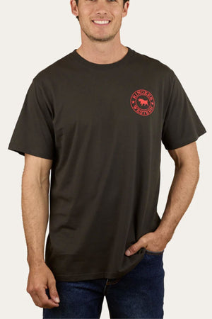 A man in a dark Ringers Western Signature Bull Loose Fit T-Shirt, made of 100% cotton with a small red circular chest graphic and text, paired with blue jeans, stands smiling with one hand in his pocket.