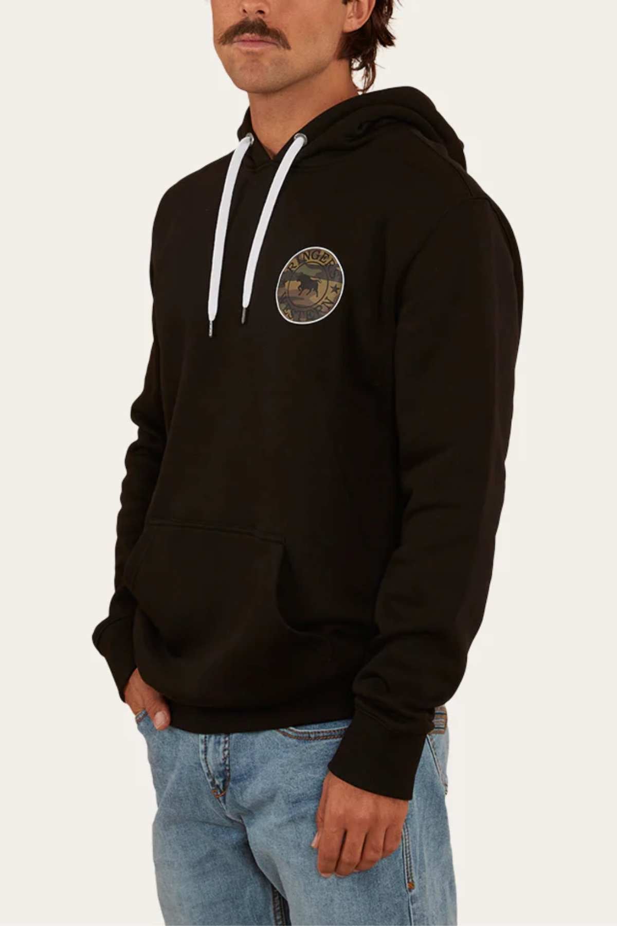 A man with a mustache is sporting a relaxed fit black Ringers Western Signature Bull Pullover Hoodie, which features a circular logo on the upper left chest. The casual hoodie has white drawstrings and a front pocket. He pairs it with blue jeans and has his right hand in the pocket. The background is plain white.