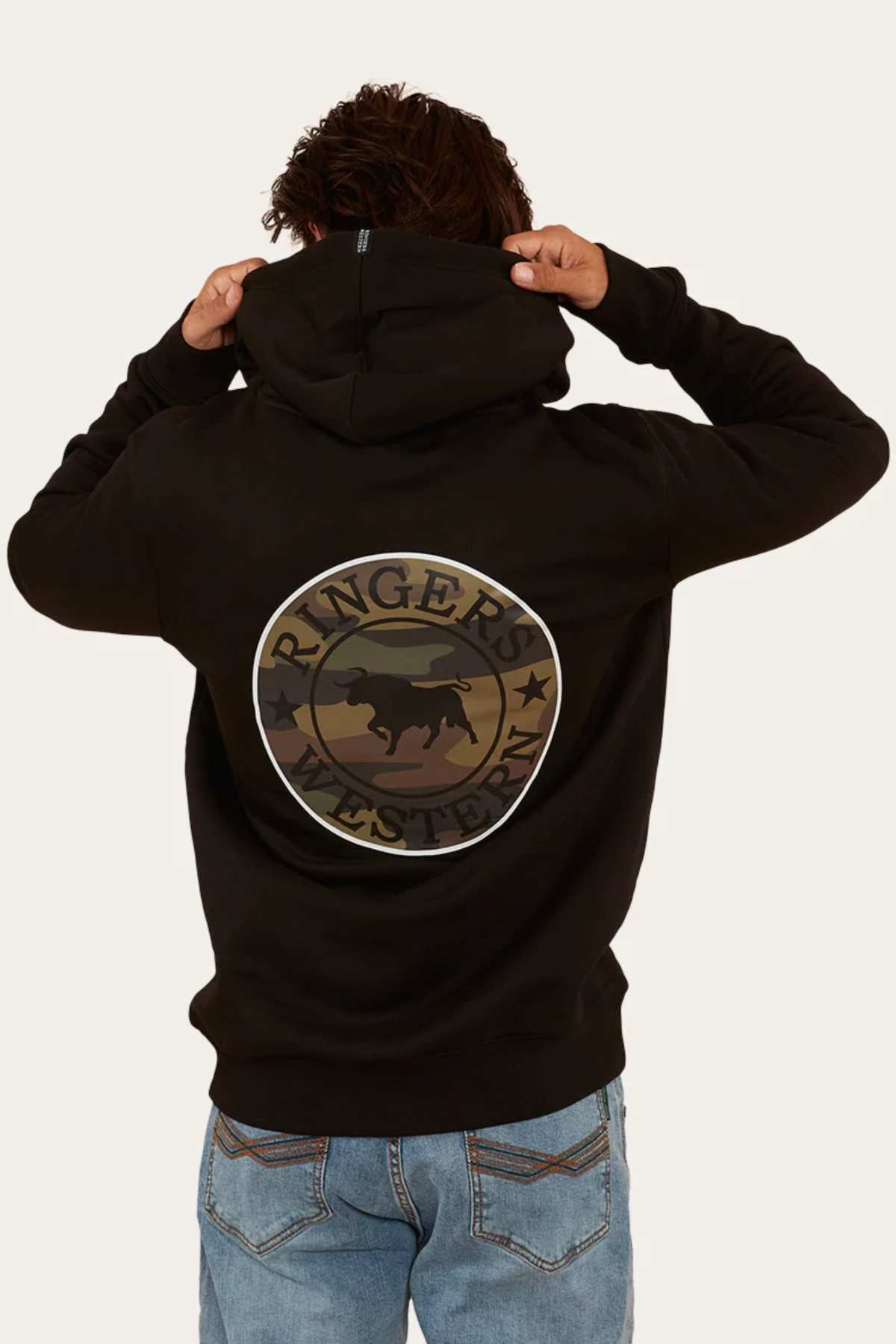 Wearing blue jeans, a person with curly hair sports the Ringers Western Signature Bull Pullover Hoodie in black, with the hood partially lifted. The hoodie features a circular logo on the back that reads "Ringers Western" encircling a silhouette of a bull. The background is plain off-white.