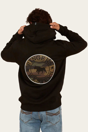 Wearing blue jeans, a person with curly hair sports the Ringers Western Signature Bull Pullover Hoodie in black, with the hood partially lifted. The hoodie features a circular logo on the back that reads "Ringers Western" encircling a silhouette of a bull. The background is plain off-white.