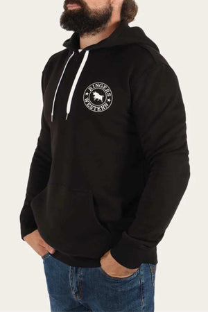 A bearded man is seen wearing a black Ringers Western Signature Bull Pullover Hoodie, characterized by its relaxed fit and white drawstrings. The hoodie features a prominent "Ringers Western" circular logo on the left chest area. He has his left hand casually tucked into the front pocket of the hoodie and is paired with blue jeans. The background is plain white.