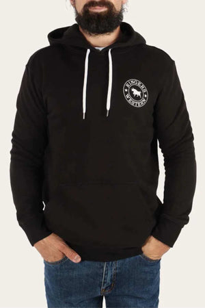 A bearded man is wearing the Ringers Western Signature Bull Pullover Hoodie, featuring a black casual design with a white drawstring and a round logo on the left chest that reads "Ringers Western" with a cow graphic in the center. He has his hands in the front pocket of the relaxed fit hoodie and is also wearing blue jeans.