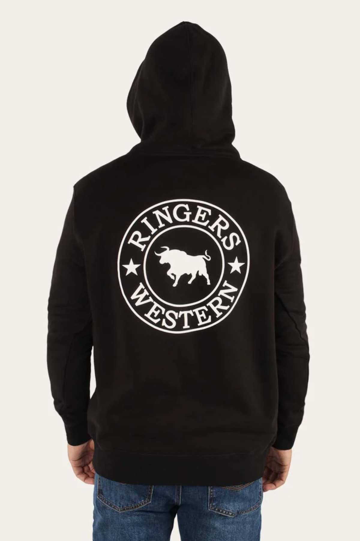 The image shows a person wearing a Ringers Western Signature Bull Pullover Hoodie in a relaxed fit with the hood up. On the back of this casual black hoodie, there's a large white circular emblem featuring "RINGERS WESTERN" around the border and a stylized bull in the center. The person pairs it effortlessly with blue jeans.