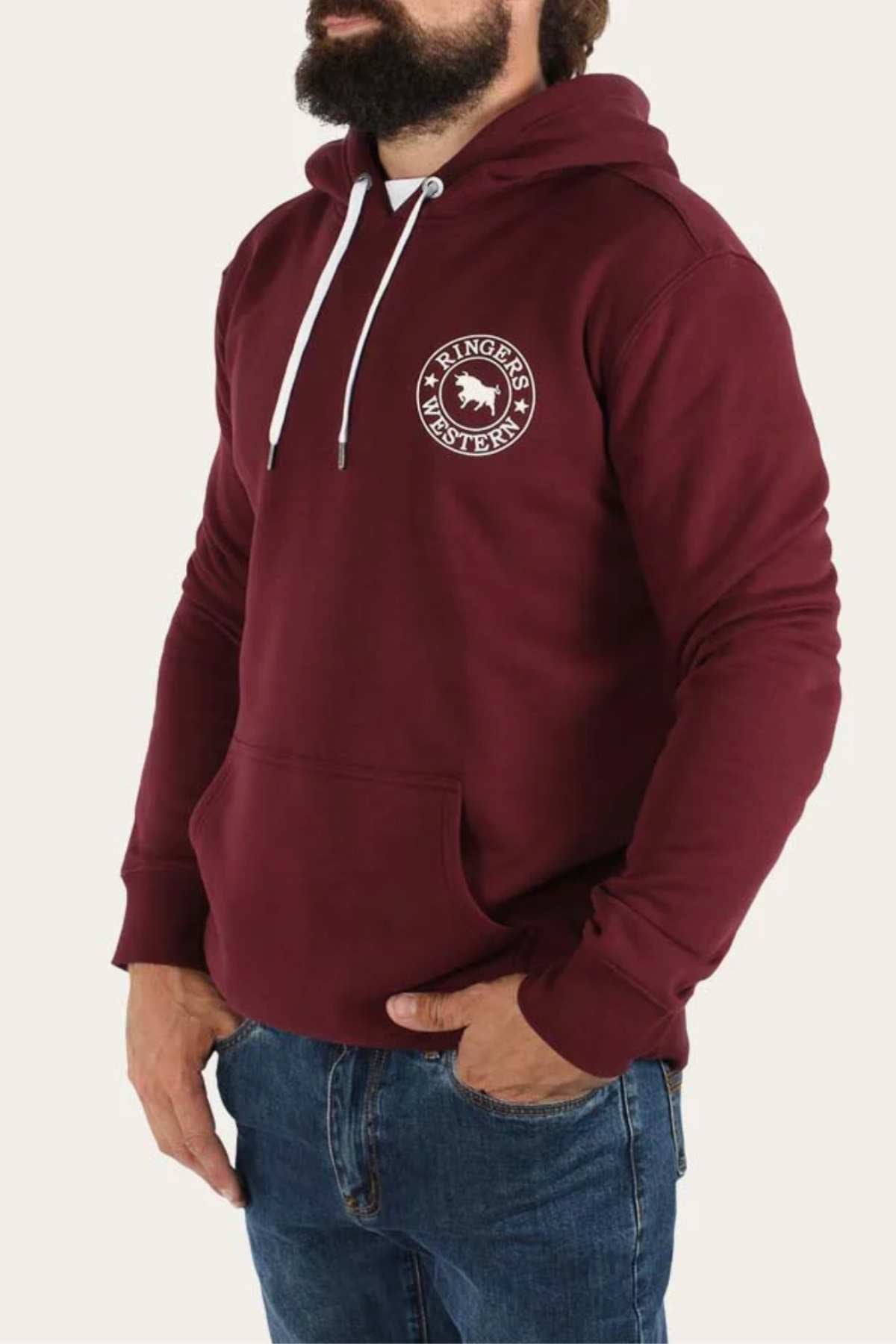 A person with a beard is wearing a maroon Ringers Western Signature Bull Pullover Hoodie, featuring a white logo on the chest and white drawstrings. This comfortable hoodie is paired with blue jeans, and they have their left hand in the hoodie pocket. The background is plain off-white.