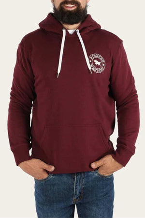 A bearded man wearing a maroon Ringers Western Signature Bull Pullover Hoodie, which features a white circular emblem with a horse and text on the left chest. The comfortable hoodie has a front pocket and white drawstrings, paired with blue jeans. He has his hands in the hoodie pocket.