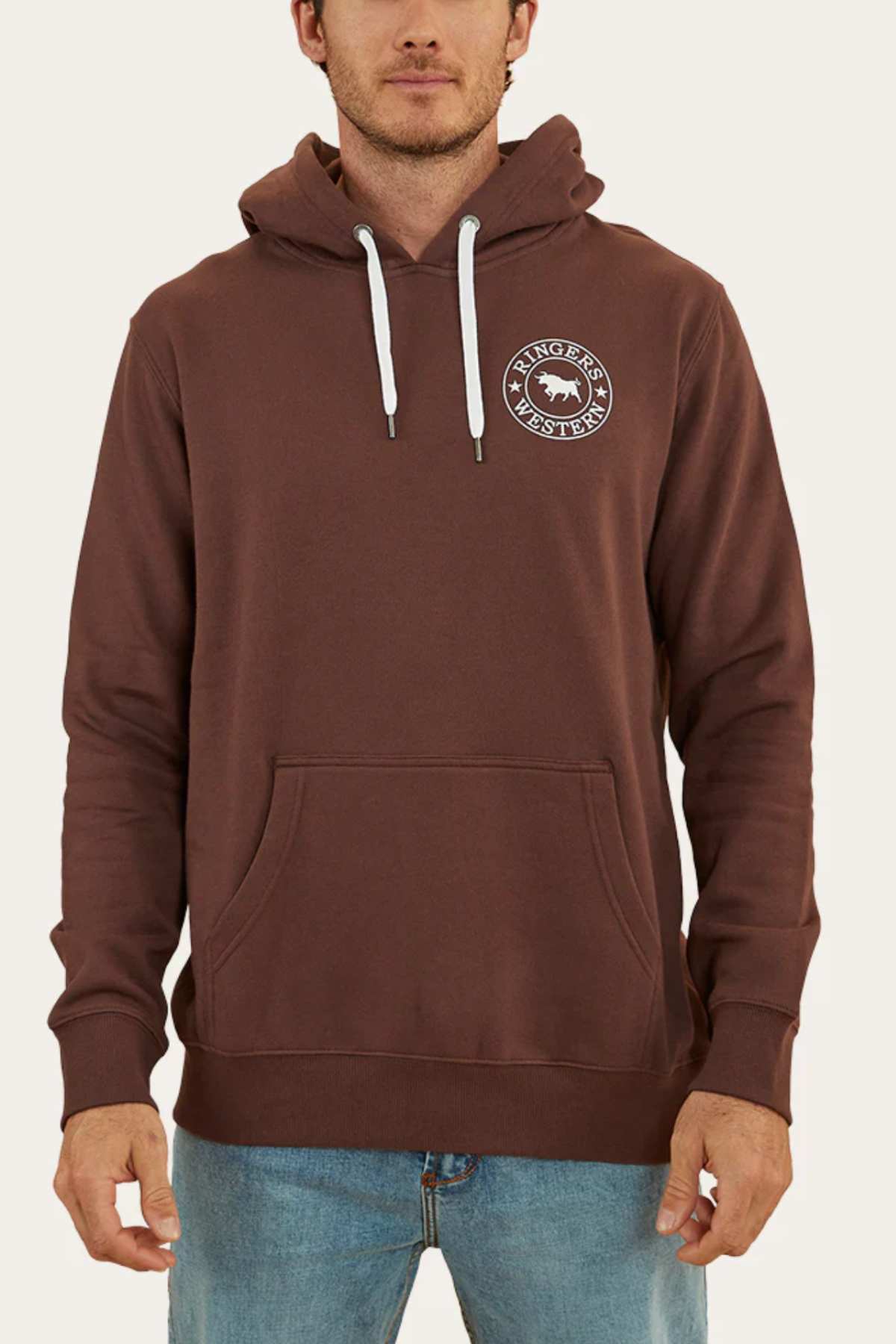 A person wearing a comfortable, brown Ringers Western Signature Bull Pullover Hoodie with white drawstrings and a circular logo on the left chest. The casual hoodie features a front kangaroo pocket, and the person is also wearing blue jeans. The background is plain and light-colored.
