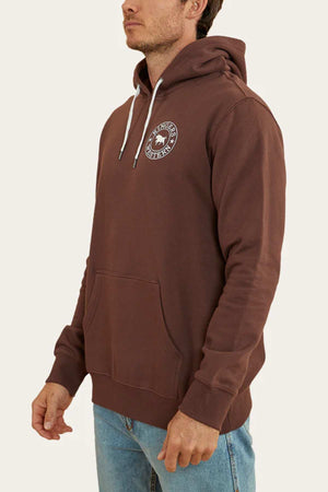 A person is wearing a comfortable Ringers Western Signature Bull Pullover Hoodie in brown, featuring a circular white logo on the left chest. The hoodie has white drawstrings and a front pouch pocket. The individual is also wearing light blue jeans and is standing sideways against a plain, light-colored background.