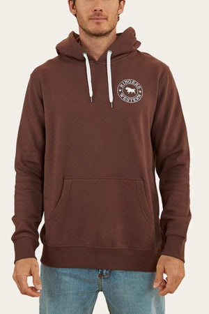 A person wearing a comfortable, brown Ringers Western Signature Bull Pullover Hoodie with white drawstrings and a circular logo on the left chest. The casual hoodie features a front kangaroo pocket, and the person is also wearing blue jeans. The background is plain and light-colored.