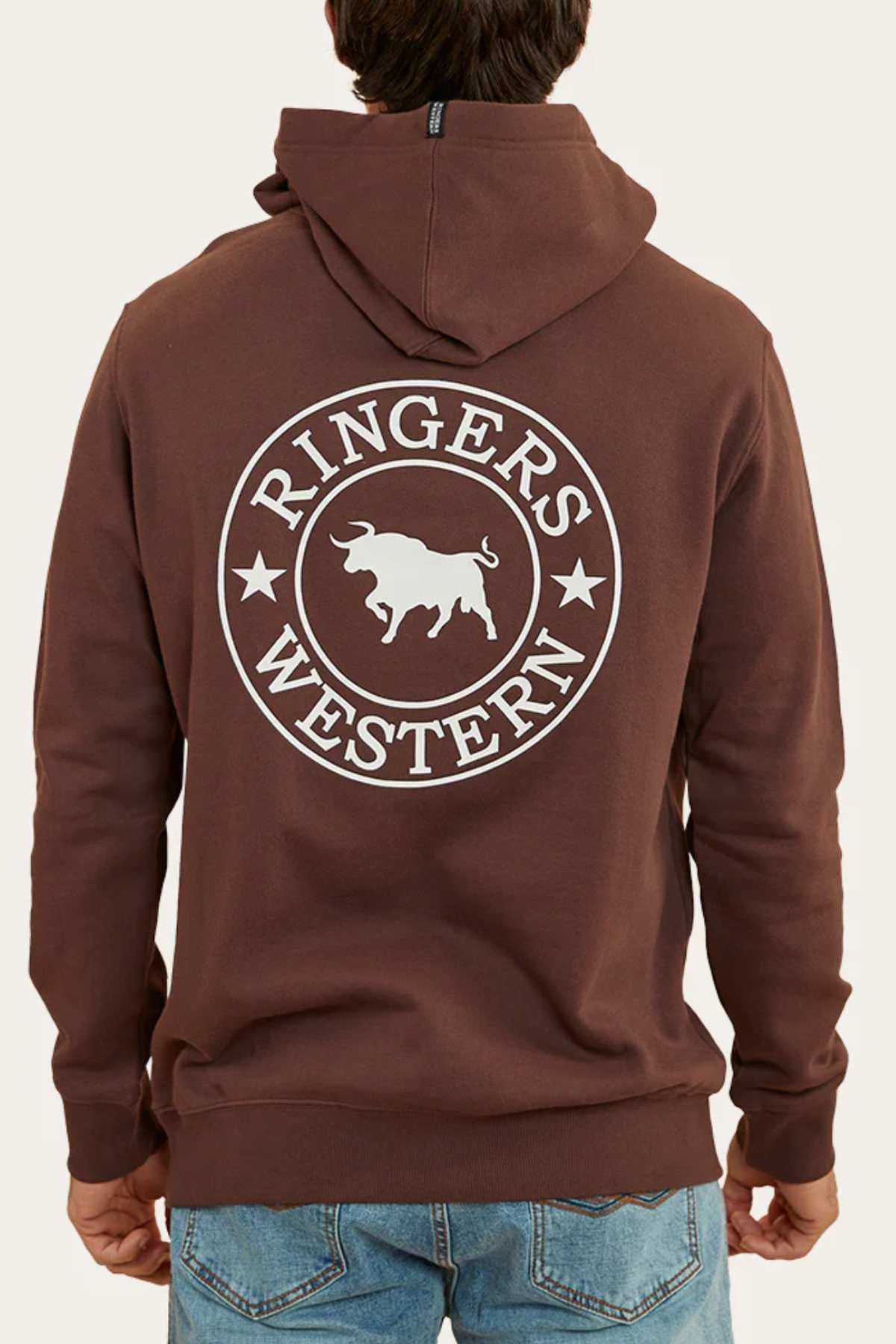 A person wearing a comfortable Ringers Western Signature Bull Pullover Hoodie in brown, featuring "Ringers Western" and a bull graphic printed on the back. The individual is standing with their back facing the camera, showcasing the design, paired with blue jeans.