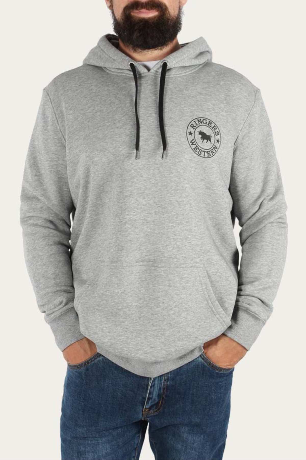 A man stands wearing a relaxed fit Ringers Western Signature Bull Pullover Hoodie in light gray with a front pocket and black drawstrings, featuring a small emblem on the left chest. He has his hands in the pockets of his blue jeans. The background is a plain off-white color.