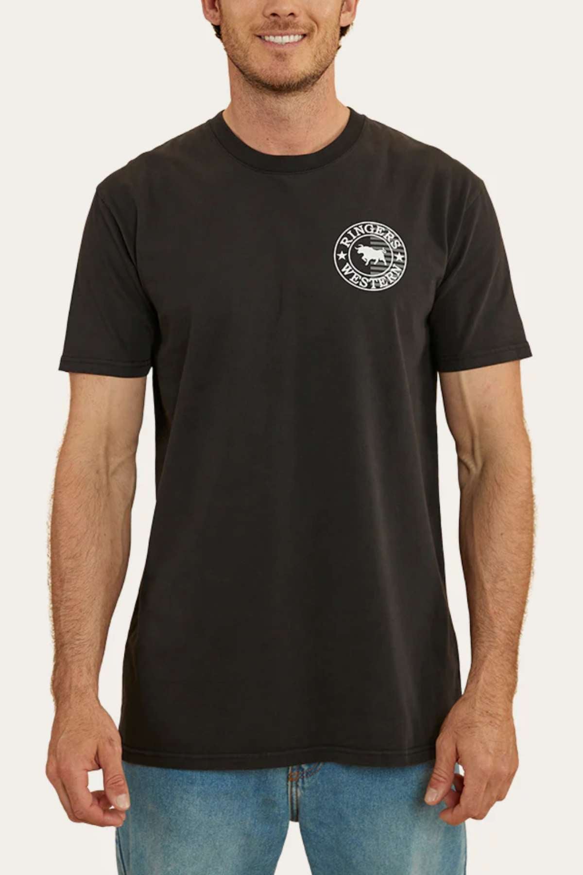 A person stands wearing a black short-sleeve Ringers Western Signature Flag Stock Fit T-Shirt crafted from 100% cotton jersey, showcasing a small, circular logo on the left chest featuring a wolf and the text "WOLVERINE RANCH" around the edge. The person is also wearing blue jeans. The background is plain white.