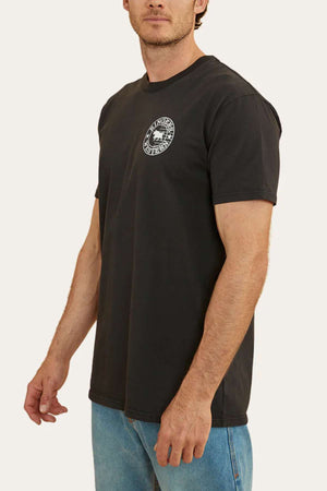 A person is wearing a black Ringers Western Signature Flag Stock Fit T-Shirt, made of 100% cotton jersey, featuring a small white circular logo on the left chest. They are also dressed in light blue jeans, standing against a plain background. The person's face is partially out of the frame.