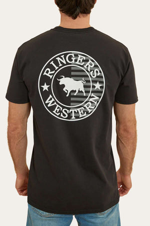 A man, clad in blue jeans and facing away from the camera, is showcasing a black Ringers Western Signature Flag Stock Fit T-Shirt made from 100% cotton jersey. The shirt features a large circular logo on the back that reads "Ringers Western" with a bull silhouette in the center, surrounded by stars and stripes.