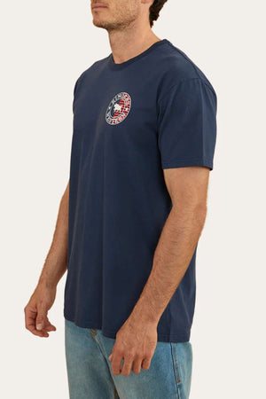 A person is standing and wearing a navy blue, 100% cotton short-sleeved Ringers Western Signature Flag Stock Fit T-Shirt with a small circular emblem on the left chest. The emblem features an American flag and the text "New England Patriots." Paired with light blue jeans, the background is plain beige.