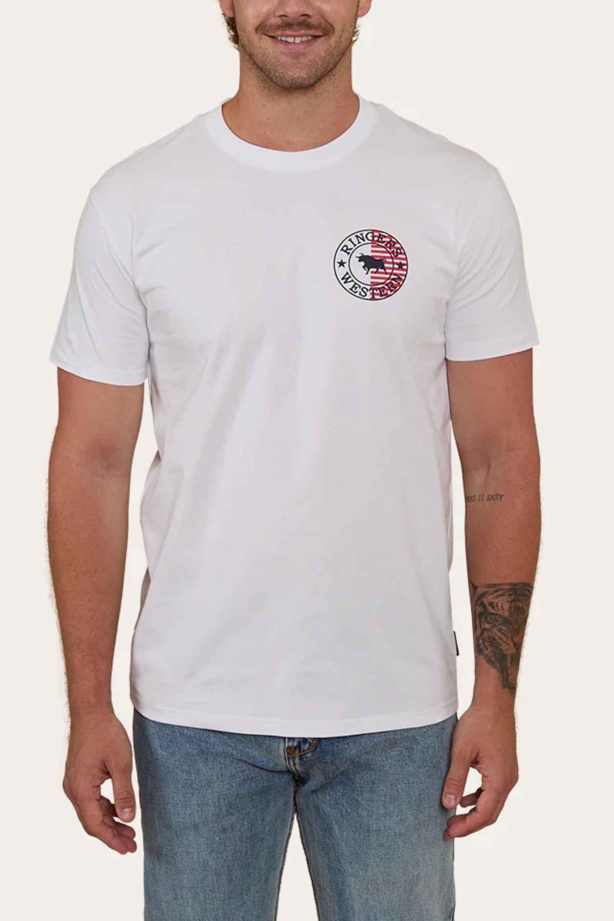 A person is dressed in a Ringers Western Signature Flag Stock Fit T-Shirt, showcasing a small circular graphic with an American flag pattern and the text "Ranger UP" on the left chest. They complement their look with blue jeans and display tattoos on their left arm.
