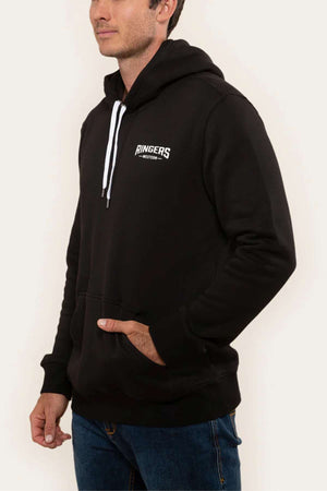 A person wearing a Ringers Western Squadron Hoodie in black, characterized by white drawstrings and a subtle white logo on the chest. The brushed fleece sweatshirt features a convenient kangaroo pocket where they have one hand tucked in, paired with blue jeans. The backdrop is plain and light-colored.