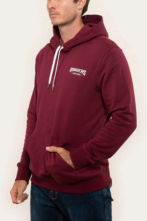 A person is wearing a maroon Ringers Western Squadron Hoodie with "RINGERS" and "WESTERN" text printed on the left side of the chest. The relaxed fit hoodie, featuring brushed fleece fabric, highlights its design against a plain, light-colored background as the individual casually rests one hand in the front pocket.