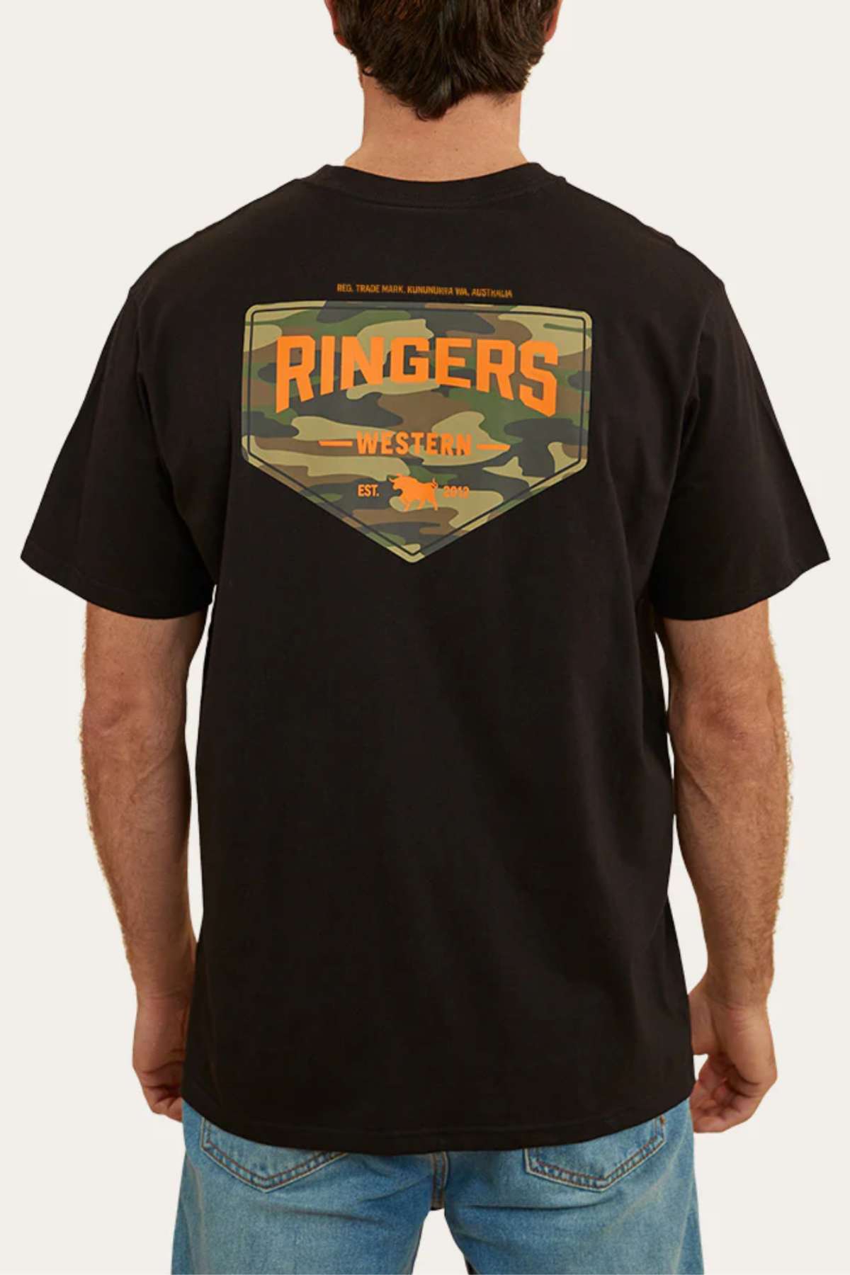 A person wearing the Ringers Western Squadron Loose Fit T-Shirt, made from pre-shrunk, 100% cotton jersey. The black t-shirt features a camo-patterned patch on the back with "Ringers Western" in large orange letters and smaller text that reads: "REAL TRADE MARK", "AUSTRALIAN 100% AUSTRALIAN", and "EST. 2012". The person