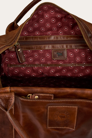 The Ringers Western Toro Gino Overniter is a brown full grain leather overnight bag with gold-tone zippers, a maroon fabric lining patterned with small logos, a zippered interior pocket, and a stitched leather logo patch inside.