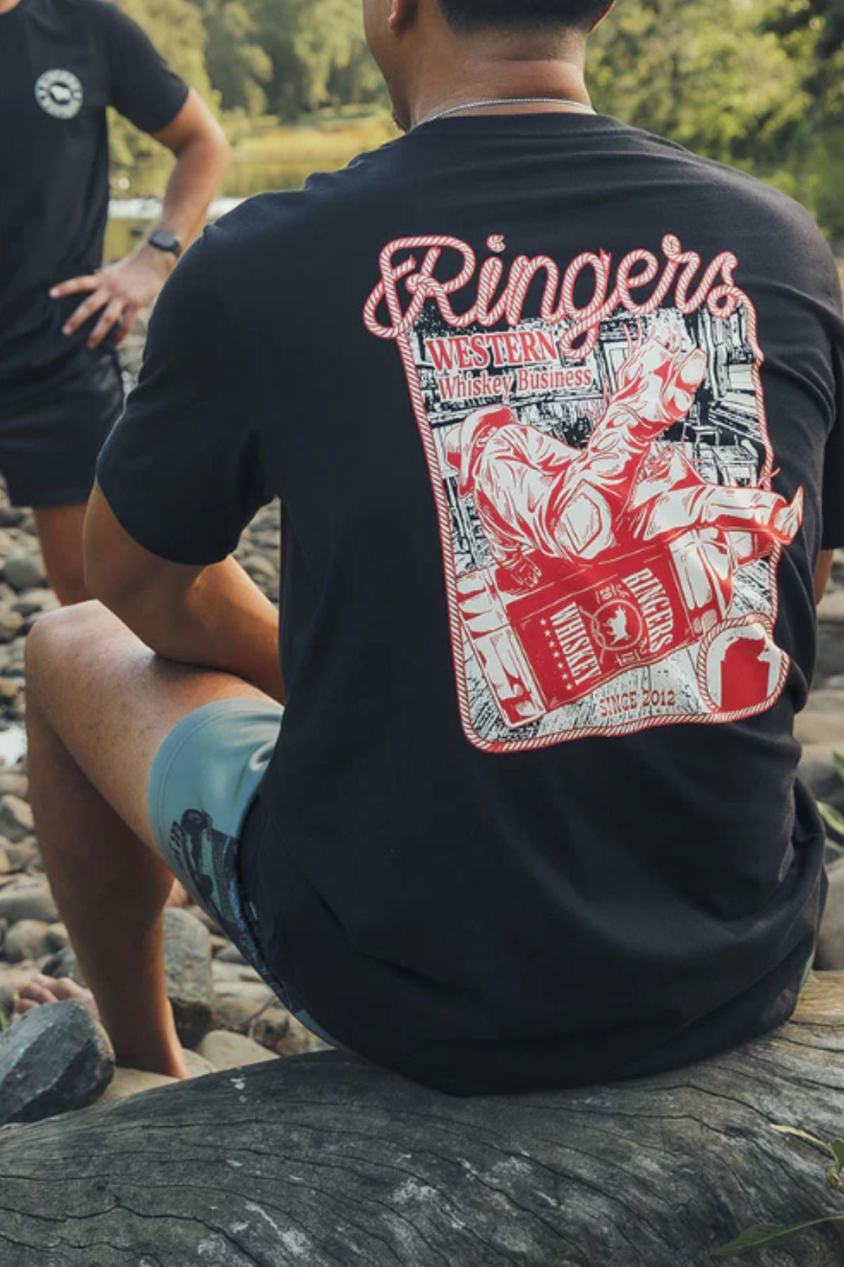 A person sits on a rocky riverside wearing a Ringers Western Whiskey Business Loose Fit T-Shirt, featuring a large red and white back graphic with a cowboy riding a bucking horse and the text βRingers Western Whiskey Business.β.