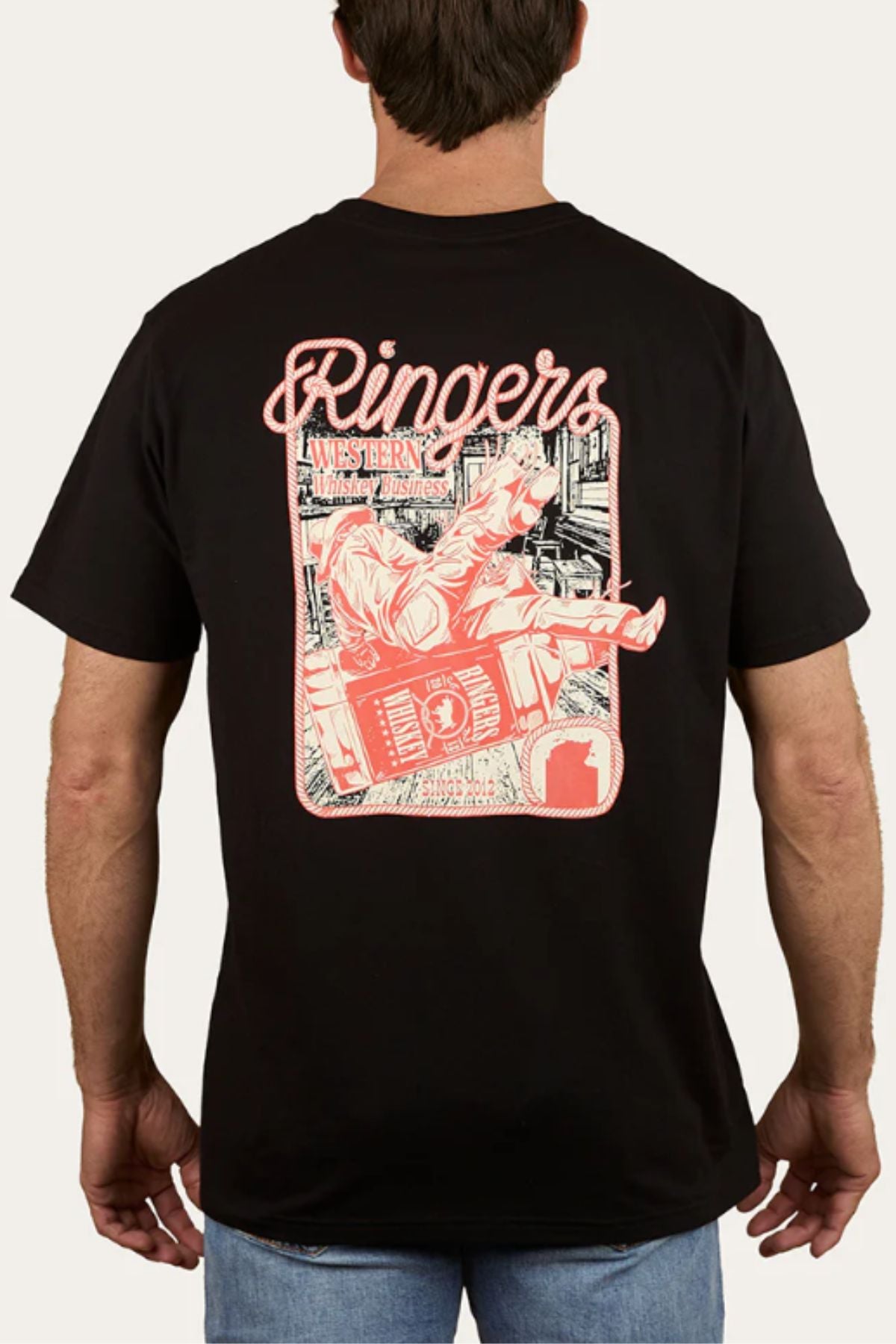 A man wears the Ringers Western Whiskey Business Loose Fit T-Shirt, featuring a bold red and white cowboy rodeo graphic on the back with city elements.